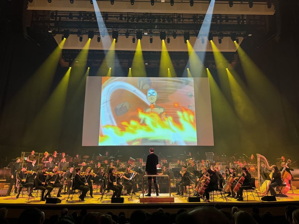 An orchestra on a stage with a still from the final battle of Avatar: The Last Airbender on a projector screen behind them.