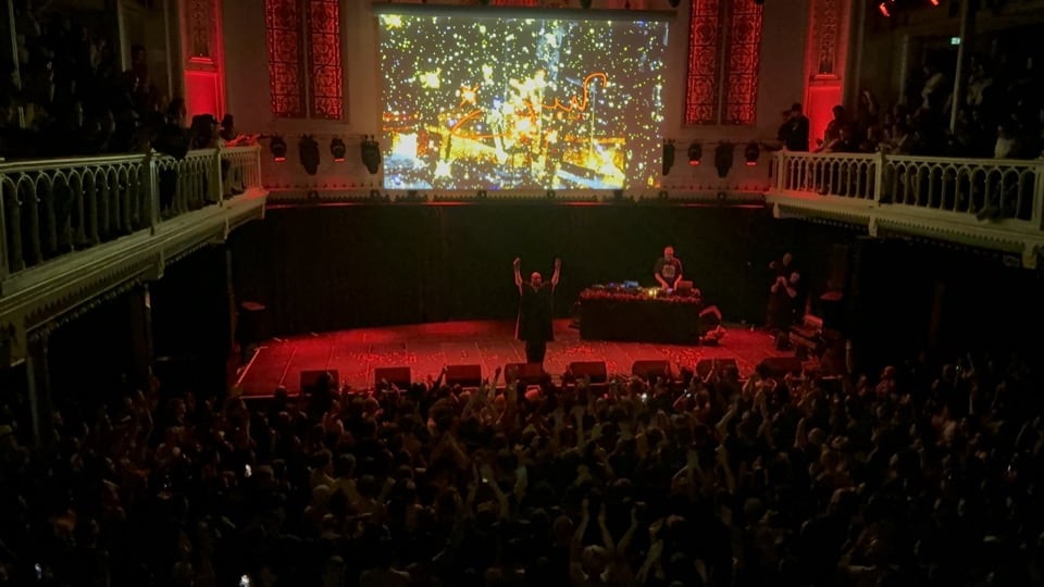 Yasiin Bey standing on stage with both hands up, facing a crowd of people. The photo was taken from above the crowd.