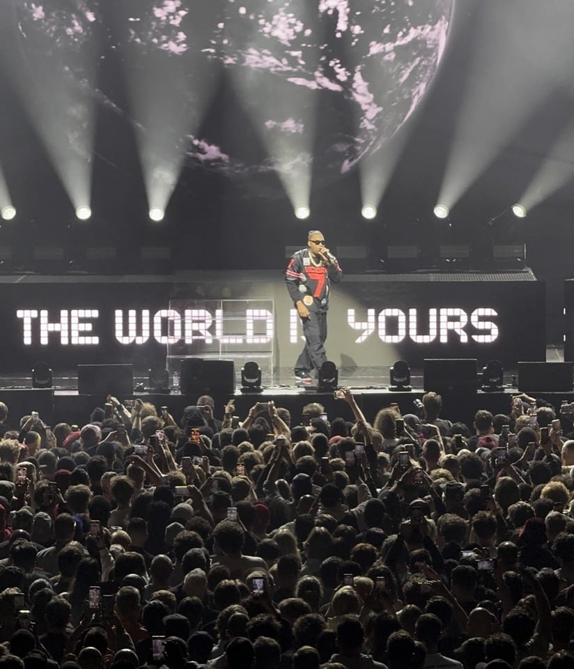 Nas performing live on stage, surrounded by the words 'The World Is Yours' on a projector behind him